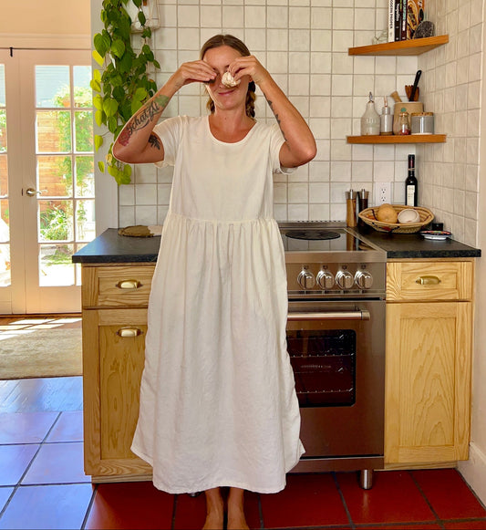 Woman in a white dress standing in a kitchen with wooden cabinets and a tiled floor.