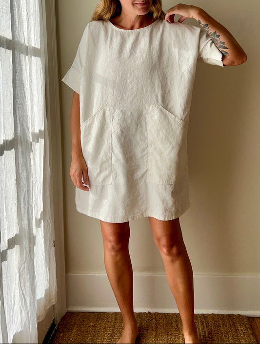 Woman wearing a white hemp tunic dress standing in a room with a window and beige wall.