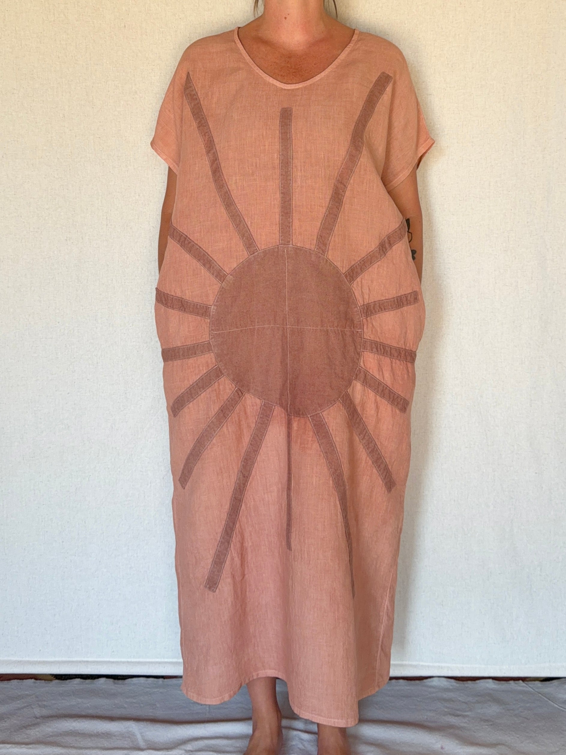 A person wearing a loose, short-sleeve midi dress in a dusty peach-pink color with a large dark brown sun motif printed or embroidered on the front, standing against a plain off-white wall.