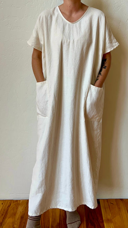 Person wearing a long white dress with pockets against a plain wall.