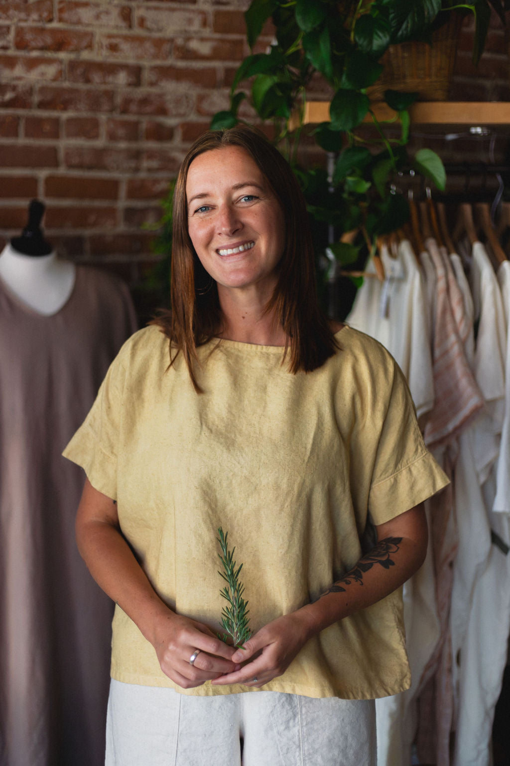 Woman holding a rosemary sprig in a clothing store with racks of clothes and a brick wall in the background.